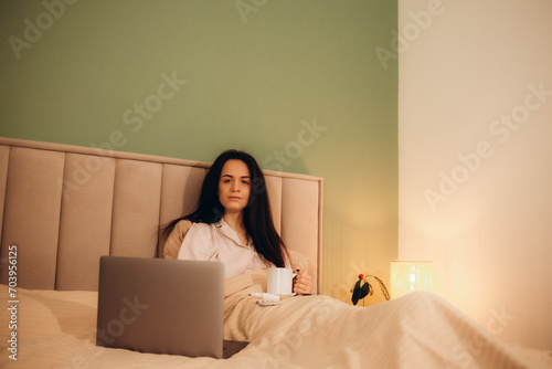 Upset sick lady in pajamas holding cup of hot drink while sitting on bed in front of modern notebook