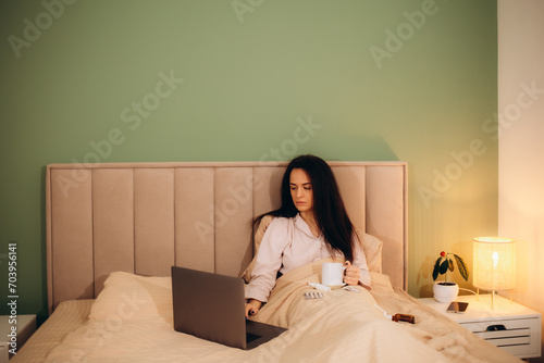 Upset sick lady in pajamas holding cup of hot drink while sitting on bed in front of modern notebook