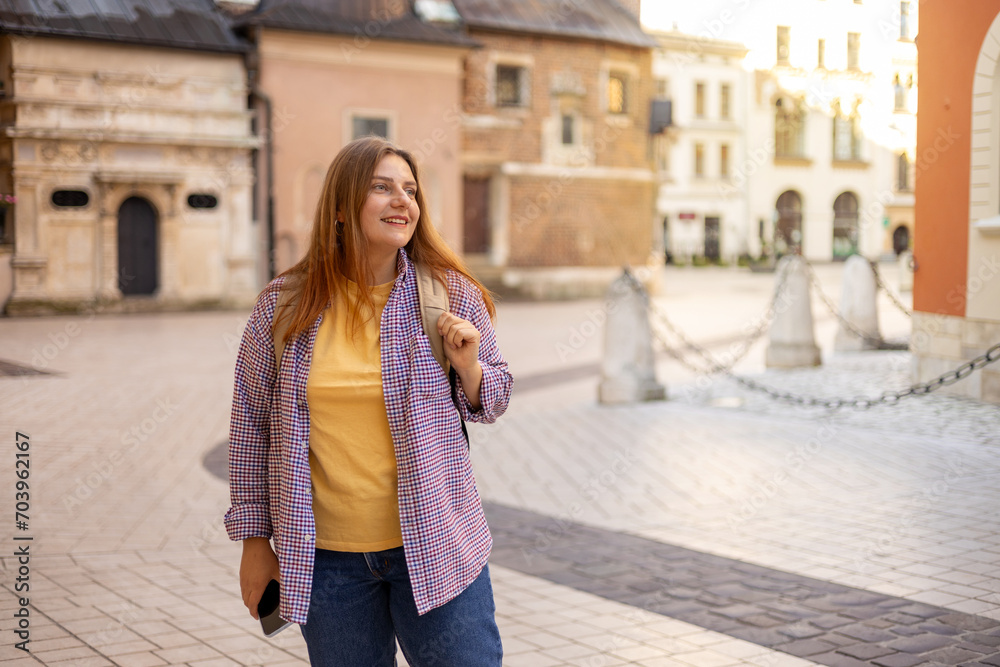 Attractive young female tourist with backpack is exploring new city. Happy 30s woman walking on old city street. Traveling Europe in summer, urban background
