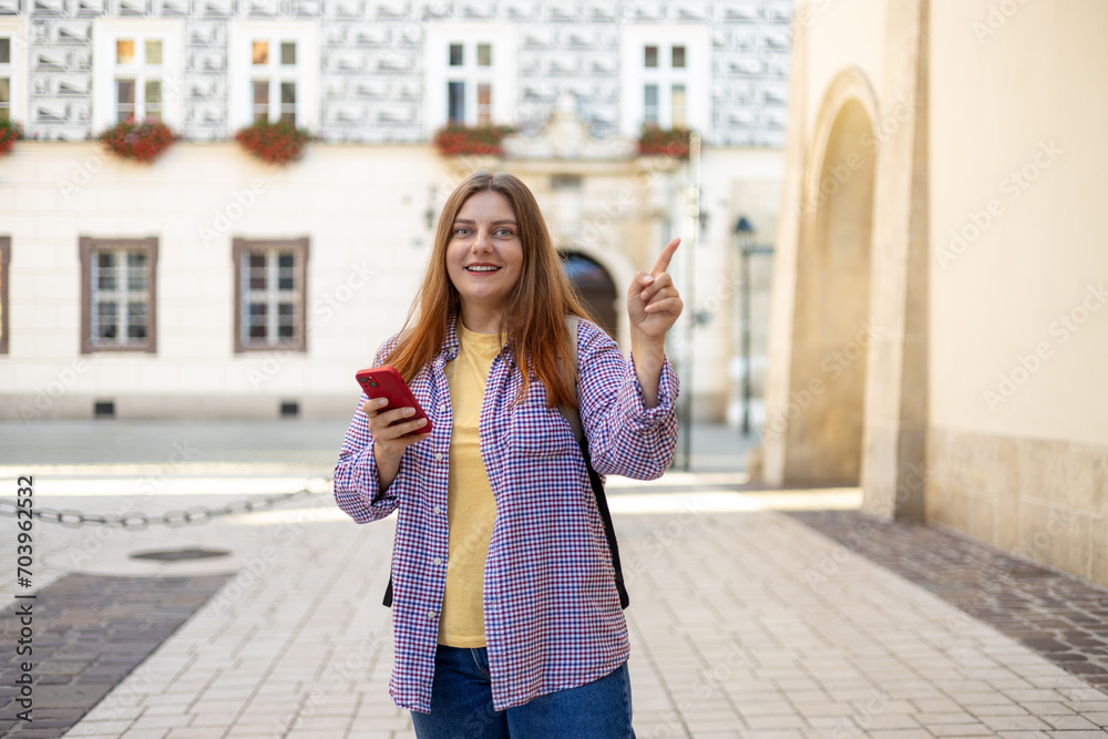 Fototapeta premium Picture of pretty young woman with backpack staying on the street. 30s tourist walking with phone and pointing finger on old city street. Use technology concept, Traveling Europe in summer