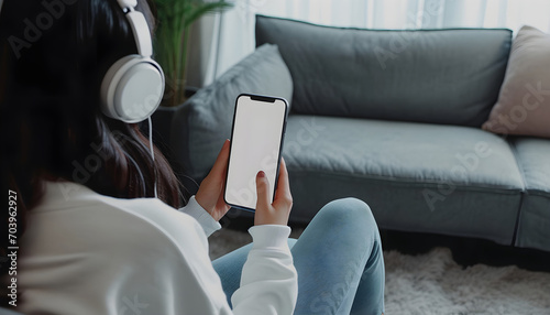 Woman with headphones holding mockup blank screen mobile smartphone while listening online music media entertainment relaxation on sofa at home, White empty space for advertise text