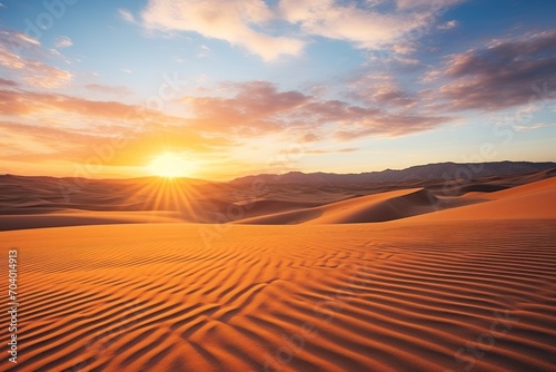  the sun is setting over a desert with sand dunes in the foreground and a mountain range in the distance, with a blue sky and white clouds in the background.