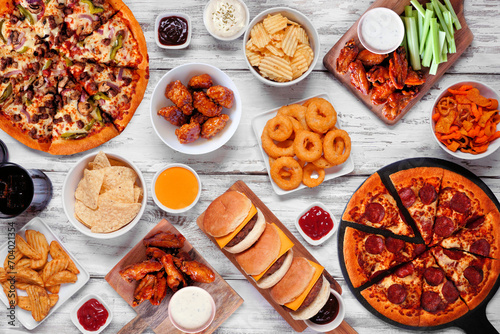 Junk food table scene. Pizza, hamburgers, chicken wings and salty snacks. Top down view over a white wood background.