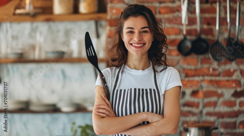 Smiling woman housewife stands with arms crossed in front of chest with spatula for cooking. Lady homemade confectioner or cook posing in striped apron draped over casual clothes photo