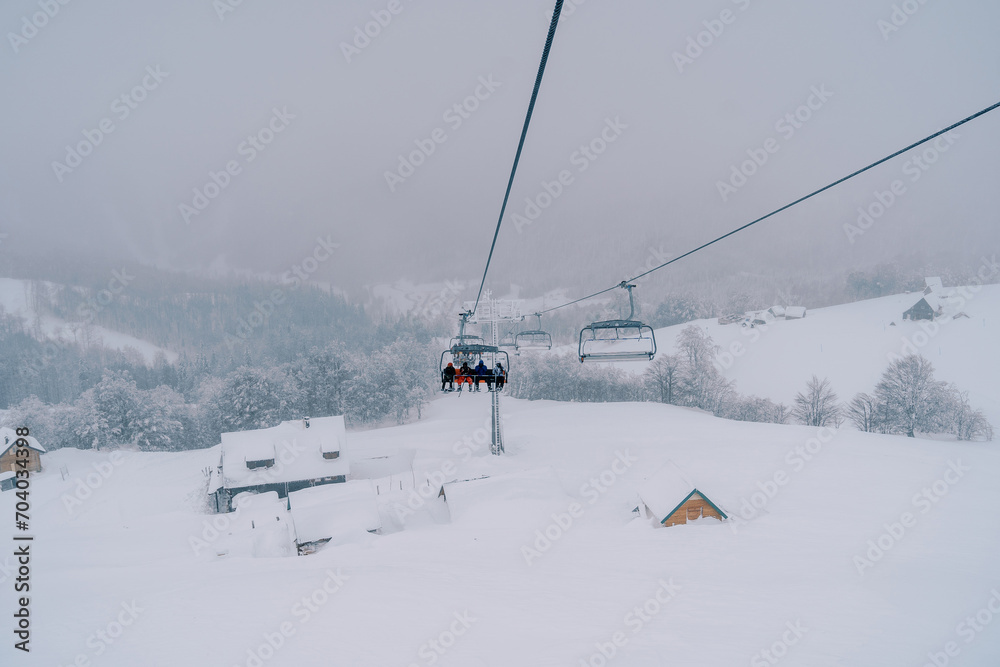 Skiers ride a chairlift up the mountain to snow-covered cottages among a coniferous forest