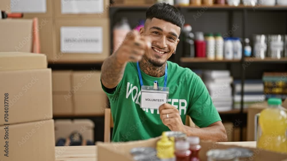 Handsome young latin man, confident in his volunteer t shirt, smiling and pointing at his identification badge then you through the camera, in the heart of charity center. Handsome young latin man, confident in his volunteer t shirt, smiling and pointing at his identification badge then you through the camera, in the heart of charity center.