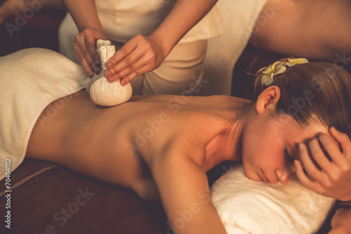 Hot herbal ball spa massage body treatment, masseur gently compresses herb bag on couple customer body. Serenity of aromatherapy recreation in warm lighting of candles at spa salon. Quiescent photo