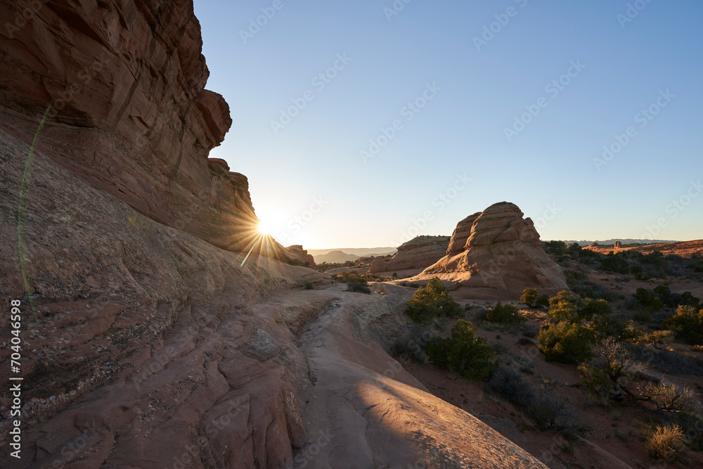 The sun peaks out behind a towering rock formation. A trail winds through the dry desert, that is sparsely populated with shrubs and greenery