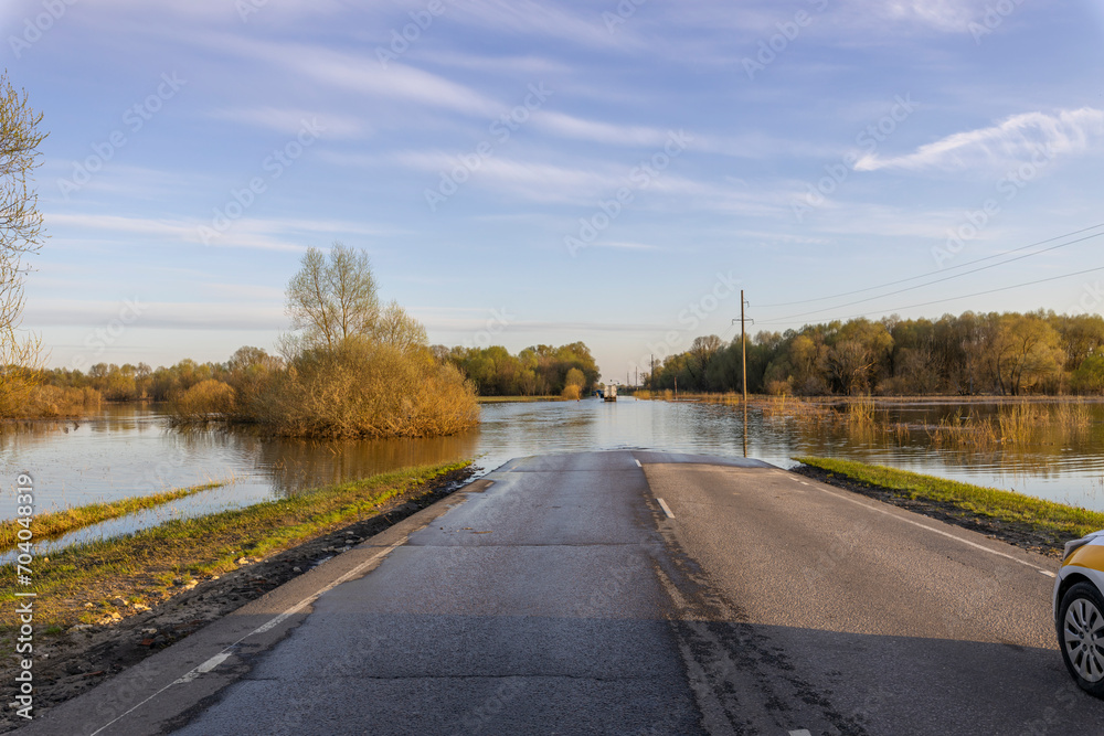Fototapeta premium The road in the countryside is flooded with water, cars drive on water, spring river flood, natural disaster, spring flood