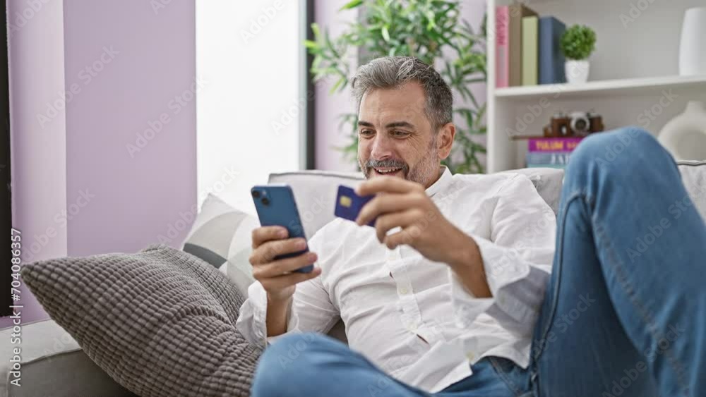 Victorious young hispanic man with a grey beard, joyfully shopping online from home, sitting on his sofa, using a smartphone to celebrate a win by making a confident investment with his credit card. Victorious young hispanic man with a grey beard, joyfully shopping online from home, sitting on his sofa, using a smartphone to celebrate a win by making a confident investment with his credit card.