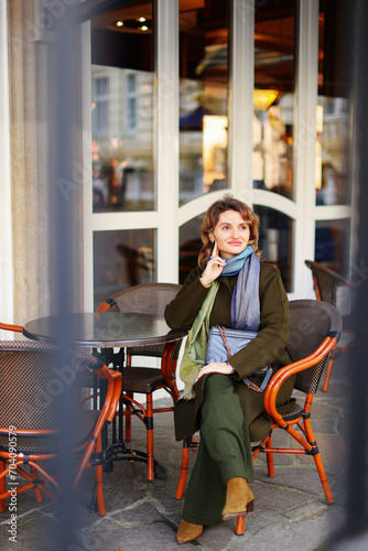 Beautiful young girl wearing green coat sitting at a table in cozy street outdoor cafe