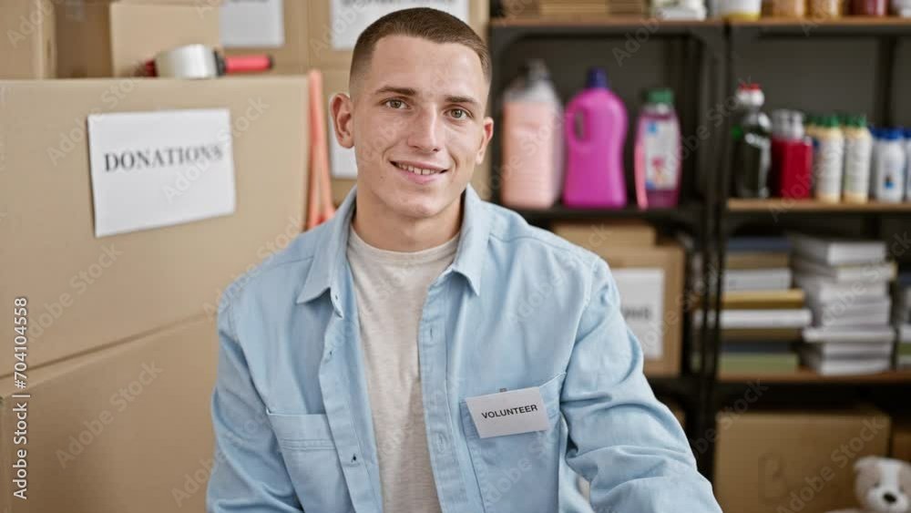 Handsome hispanic man wearing a volunteer badge poses at a charity donation center with cardboard boxes and supplies in the background.