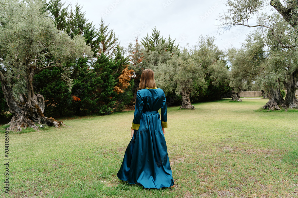 A woman in a teal dress stands contemplative among ancient olive trees in a peaceful grove A woman in a teal dress stands contemplative among ancient olive trees in a peaceful grove