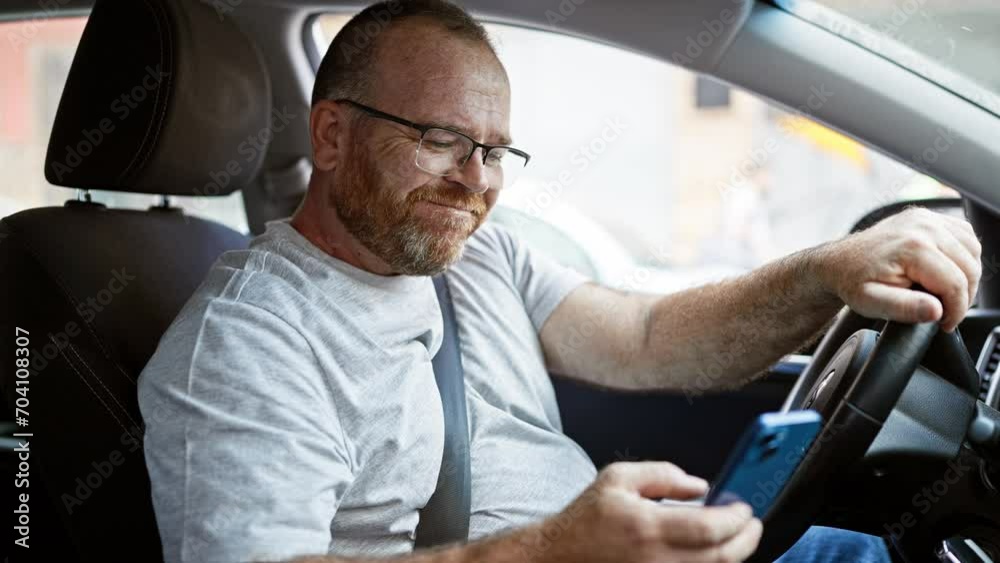 Handsome middle-aged caucasian man sitting in his car, smiling confidently as he types a message on his smartphone under the warm city sunlight
