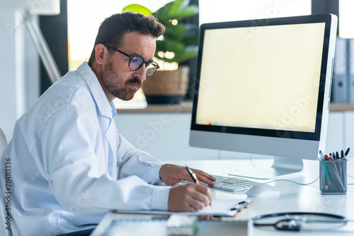 Mature male doctor working with computer while writing notes in the medical consultation