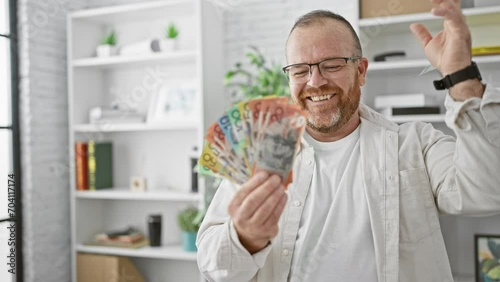 Cheerful caucasian man, handsome business worker joyfully throwing australian dollars in air at workplace, bubbling over with success, happiness, and confidence in office photo