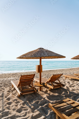 Fototapeta Naklejka Na Ścianę i Meble -  Wooden loungers await under rustic straw umbrellas on a sun-kissed beach, relaxation by the turquoise sea