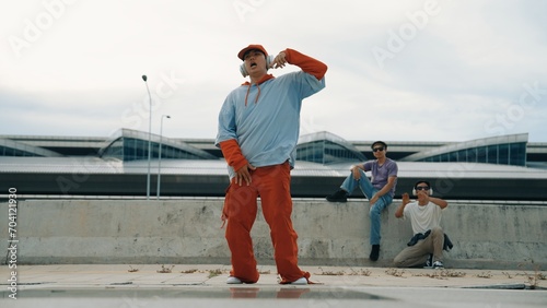 Hip hop man wearing headphone while pose at camera with confident at bridge. Active energetic hipster perform freestyle dance or footstep while friend cheering behind. Outdoor music 2024. Sprightly.