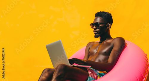 An African American man with sunglasses and a swim ring uses a laptop against a vibrant yellow background, capturing the essence of travel and summer