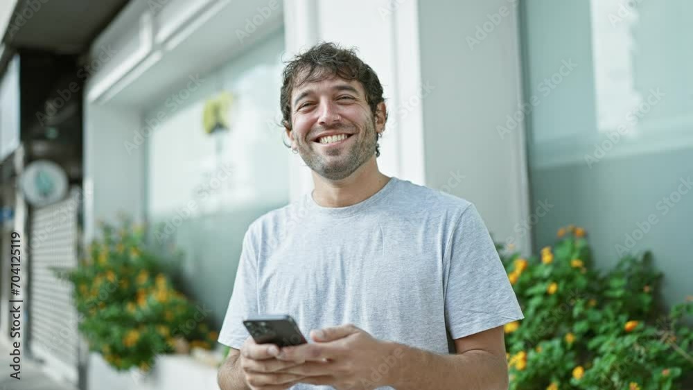 Happy young man, sporting a beard and blond hair, enjoying his time outdoor in the city, confidently smiling while typing a message on his smartphone screen Happy young man, sporting a beard and blond hair, enjoying his time outdoor in the city, confidently smiling while typing a message on his smartphone screen