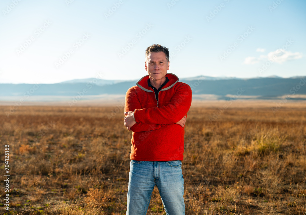 Middle aged man farmer, rancher, real estate developer, CEO, in orange fleece pullover standing on vast high plains grassland with Rocky Mountains in the background Middle aged man farmer, rancher, real estate developer, CEO, in orange fleece pullover standing on vast high plains grassland with Rocky Mountains in the background