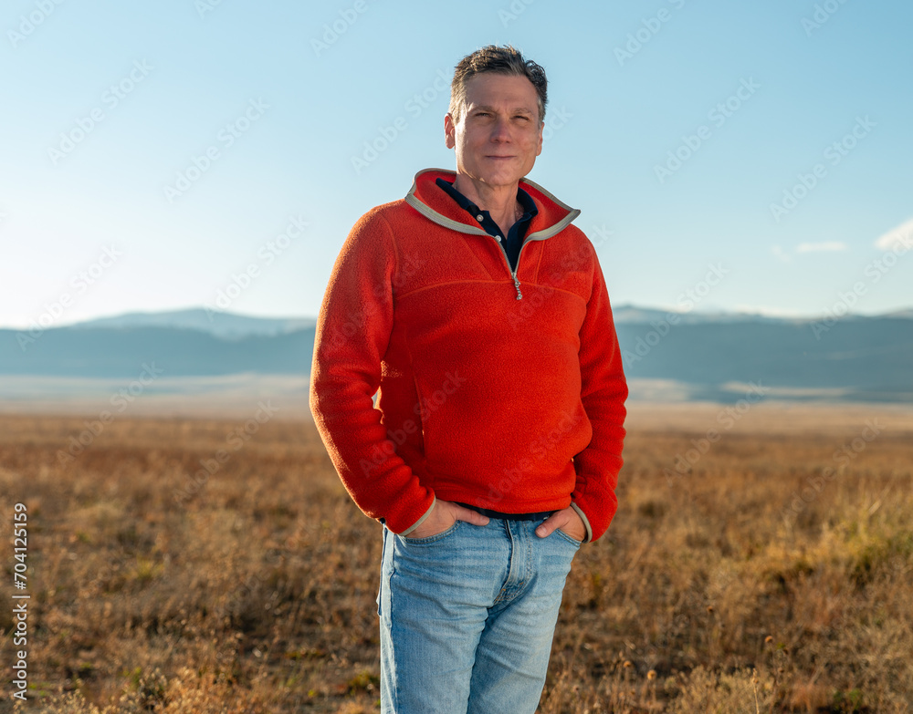 Middle aged man farmer, rancher, real estate developer, CEO, in orange fleece pullover standing on vast high plains grassland with Rocky Mountains in the background