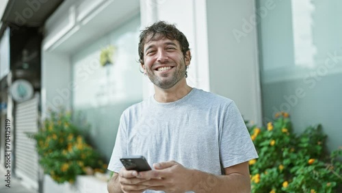 Happy young man, sporting a beard and blond hair, enjoying his time outdoor in the city, confidently smiling while typing a message on his smartphone screen photo