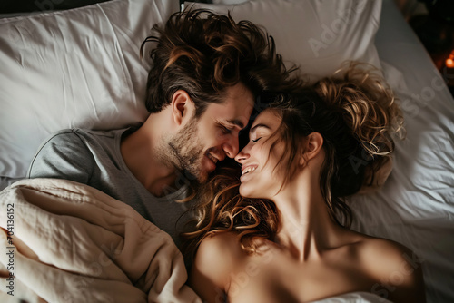 Young beautiful and loving couple wake up while lying on white pillows under the covers on soft white bed at the morning photo