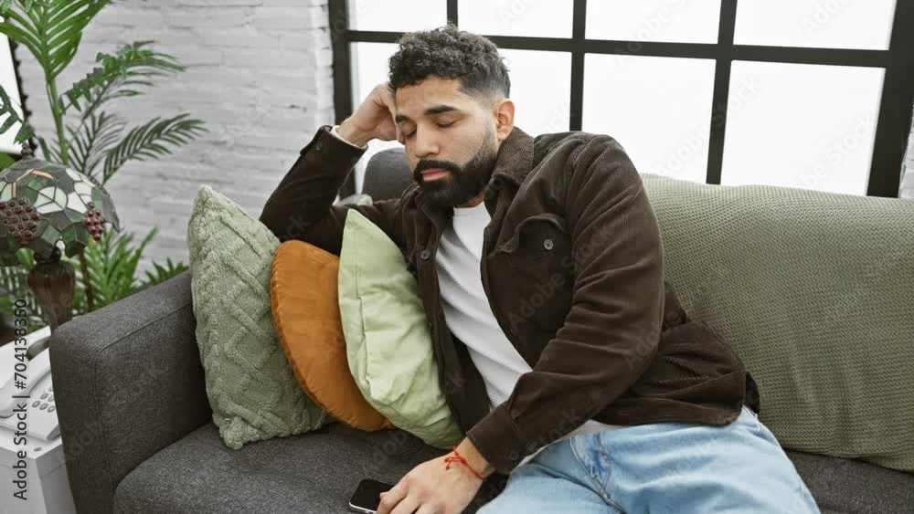 A pensive man with a beard sits on a couch indoors, holding a phone in a stylish living room. A pensive man with a beard sits on a couch indoors, holding a phone in a stylish living room.
