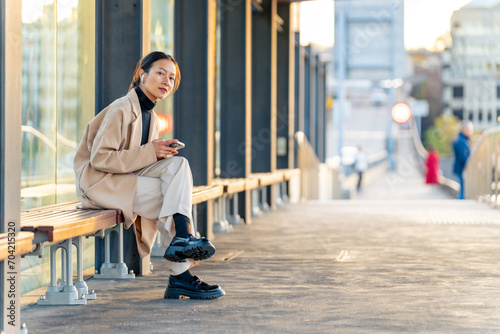 Happy Asian woman using mobile phone with mobile app chatting or social media during waiting for tram at station. Attractive girl enjoy urban outdoor lifestyle travel city street with smartphone.