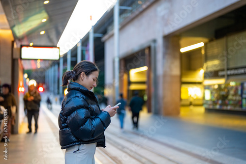 Happy Asian woman using mobile phone with mobile app chatting or social media during waiting for tram at station. Attractive girl enjoy urban outdoor lifestyle travel city street with smartphone. photo