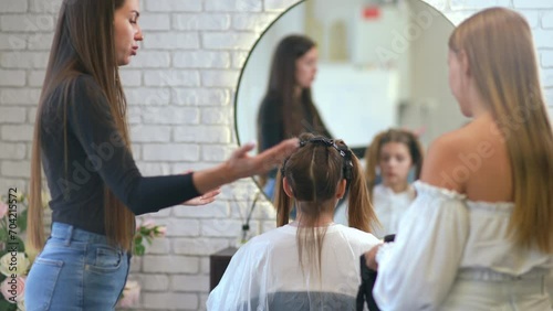 Female hairdresser mentor explains hair coloring procedure while newbie woman puts on black gloves standing behind female client in hair salon. photo