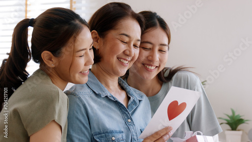 Mother day two grown up child cuddle hug give flower gift box red heart card to mature mum. Love kiss care mom asia middle age adult three people sitting at home sofa happy smile enjoy family time. photo