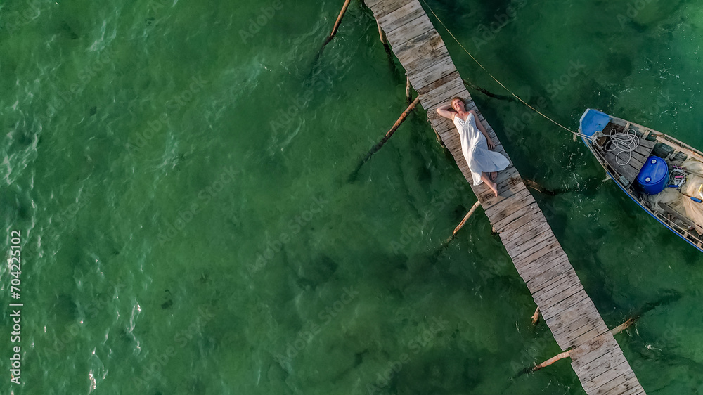 Aerial view of a person relaxing on a rustic wooden pier over clear green waters next to a small boat, depicting tranquility or a tropical vacation concept