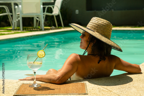 Mulher morena com chapéu de palha de costas curtindo o sol na beirada da piscina e ao seu lado um refrescante drink de limão. photo