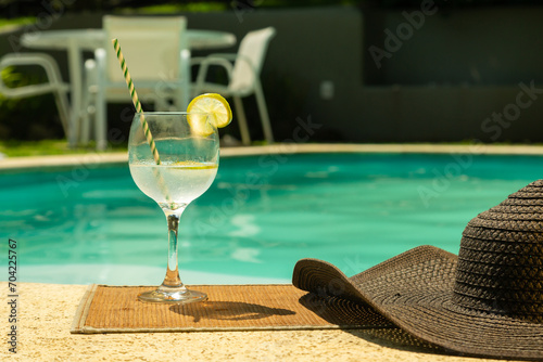 uma taça de vinho de vidro transparente, cheia de vinho branco, com um canudo de papel e uma fatia de limão, ao lado um chapéu de sol
em um dia ensolarado e ao fundo uma piscina azul. photo