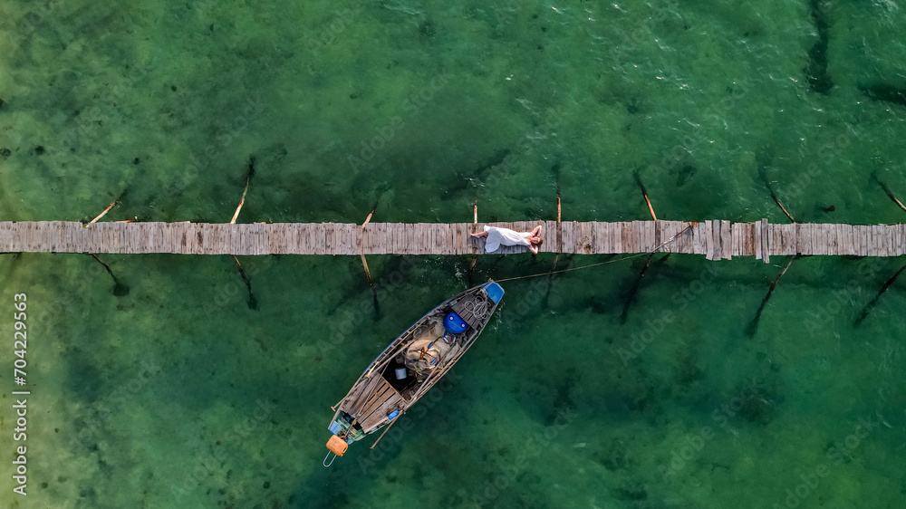 Aerial view of a person standing on a narrow wooden jetty above clear green water with a wooden boat below, depicting travel or leisure concept