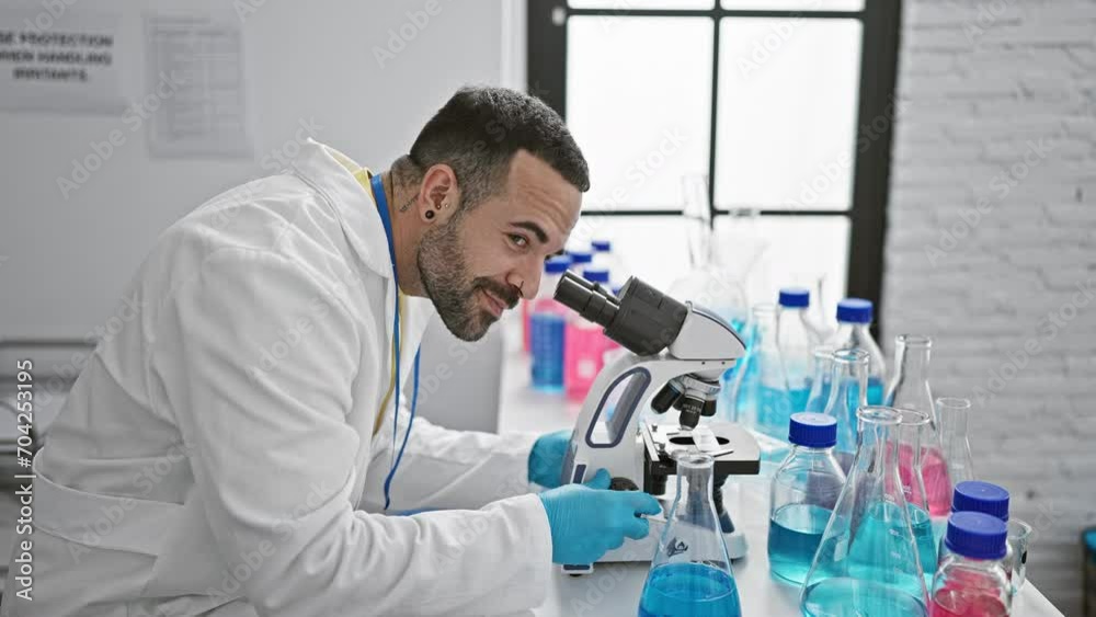 Handsome hispanic man using microscope in modern laboratory setting with blue gloves and beakers around. Handsome hispanic man using microscope in modern laboratory setting with blue gloves and beakers around.
