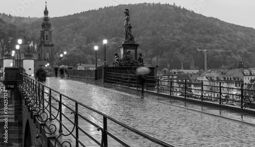 Black and white photo of the old bridge (Karl-Theodor-Brücke) in Heidelberg during a rainy day. Some pedestrians are walking with umbrellas. Blurred movement effect of the people on the bridge. #704257174