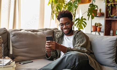 Smiling young African American guy sitting on couch using phone. Happy relaxed gen z teen at home holding cellphone chatting in mobile apps on cell phone in modern living room, Generative AI  photo