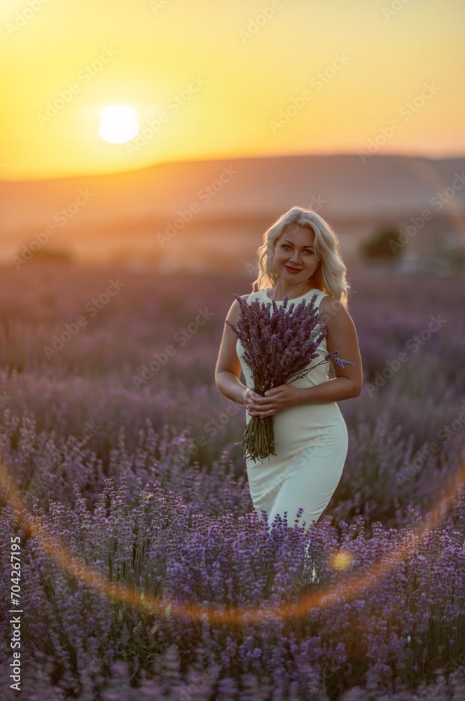 Blonde woman poses in lavender field at sunset. Happy woman in white dress holds lavender bouquet. Aromatherapy concept, lavender oil, photo session in lavender