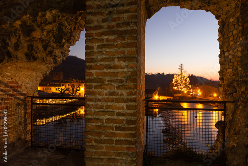 Magical Archway: A lit Christmas tree reflects in tranquil waters through a weathered brick arch. Warm-lit buildings enhance the festive ambiance. The sunset transitions, casting a soft glow