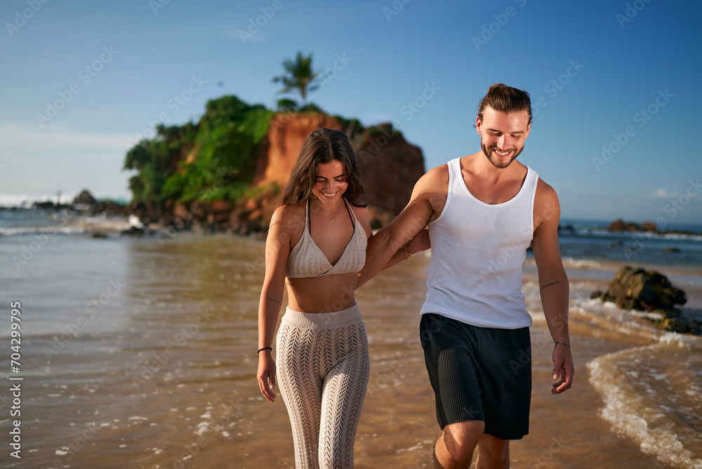 Happy couple walks barefoot on tropical beach at sunset, man leads woman by hand near ocean, romantic stroll by sea, young love on summer holiday, relaxed lifestyle, travel, vacation mood. Happy couple walks barefoot on tropical beach at sunset, man leads woman by hand near ocean, romantic stroll by sea, young love on summer holiday, relaxed lifestyle, travel, vacation mood.