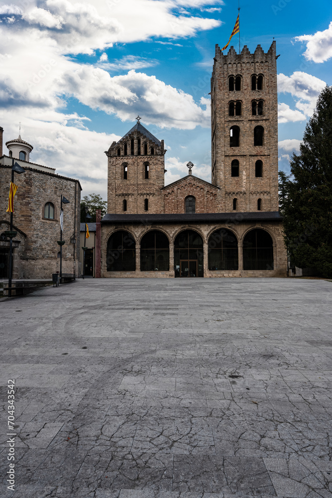 view of the Monastery of Ripoll, one of the most important and impressive monuments of Catalan Romanesque and Gothic art. The image shows the intricate portal, the towers, the dome, and the flag of