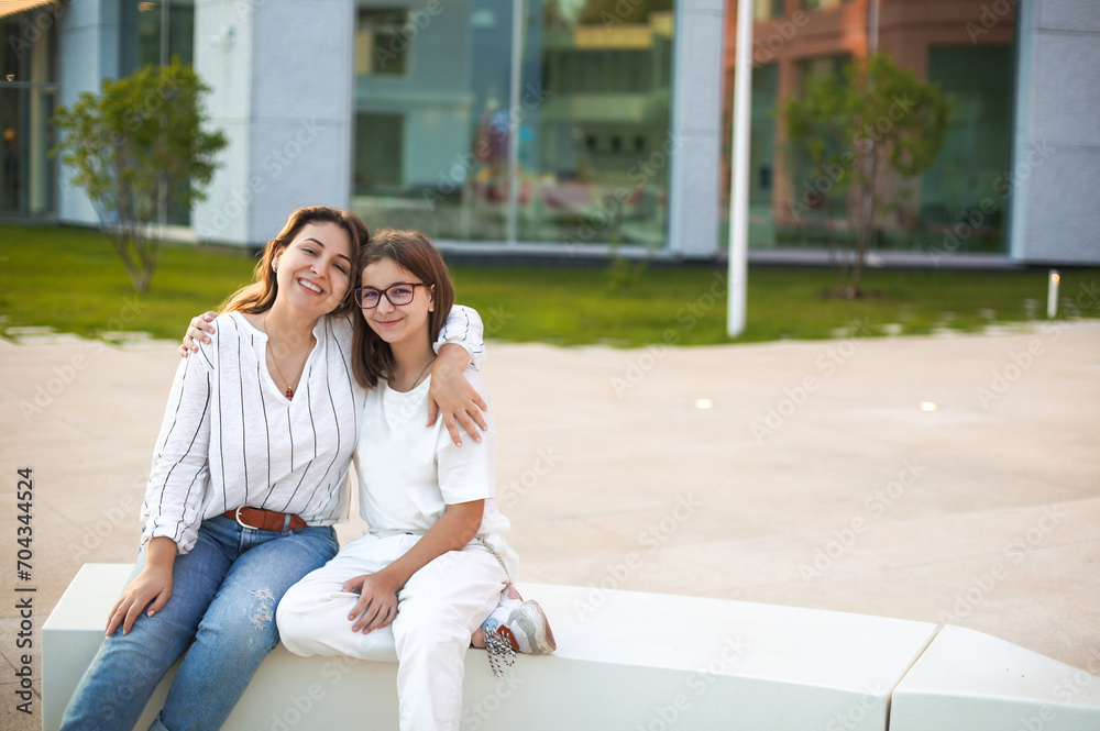 Mother and teen daughter sitting on bench on street