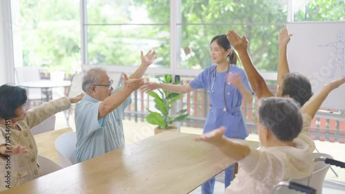 Side view young Asian nurse stand in front of group of senior or elderly people to show as example for exercise hands in room of clinic or healthcare center.