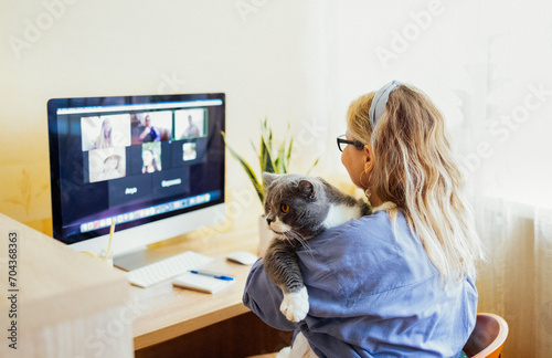 Young woman sits in front of computer monitor and talks via video link. Fair haired girl with cute gray white cat in her arms communicates on video link.