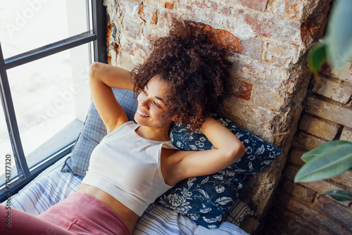 Pleased relaxed African American ethnicity girl in homewear sitting on windowsill photo