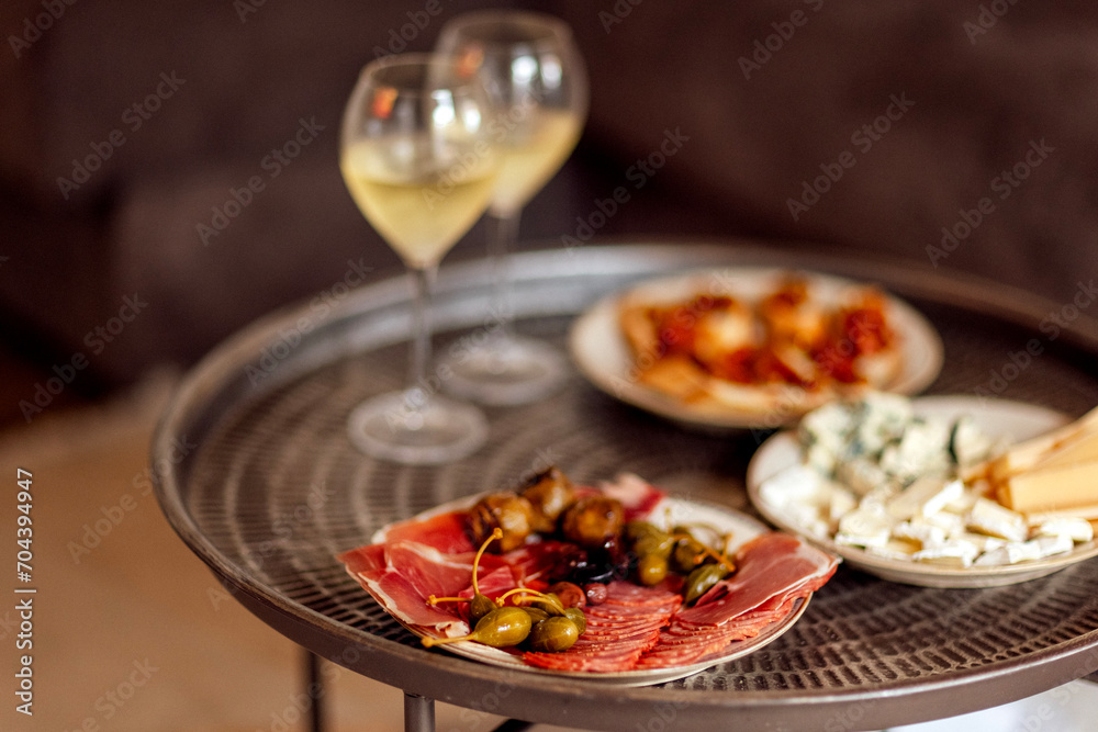 Two glasses of cool champagne and plates with various snacks on a round metal coffee table. White plates with slices of ham, salami, capers, olives and cheese.