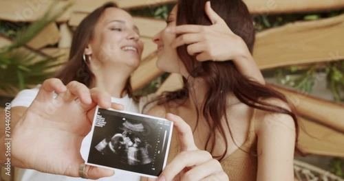 Happy lesbian women in love, expect baby and showing ultrasound picture while sitting embracing. Joyful pregnant female in same sex marriage. Maternity concept. LGBT relationship photo